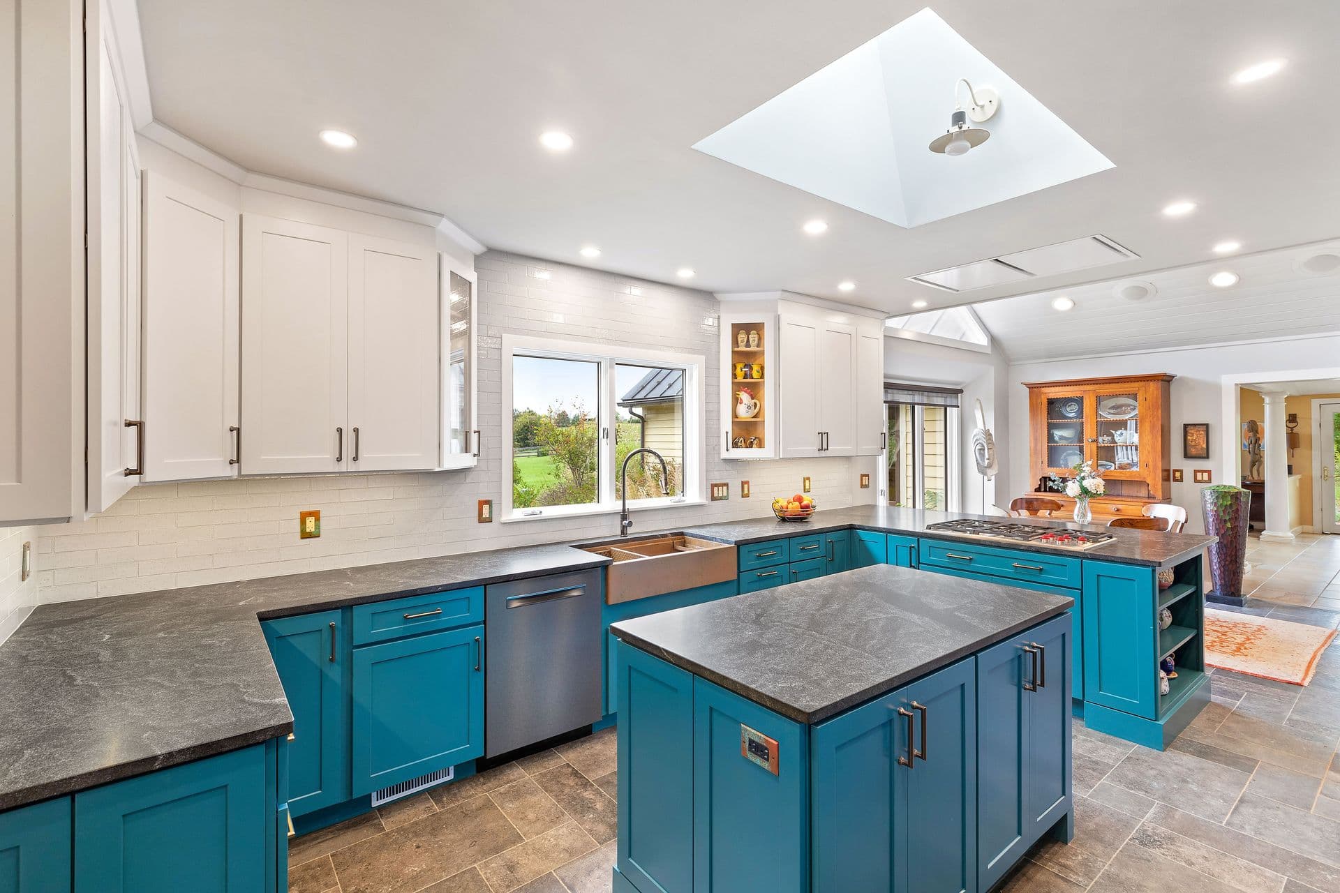 Modern kitchen with teal island and base cabinets, white upper cabinets, gray countertops, farmhouse sink, skylight, and open layout connecting to dining and living areas